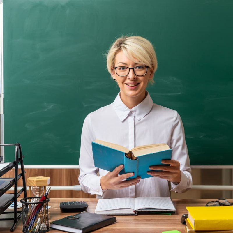 smiling-young-blonde-female-teacher-wearing-glasses-sitting-desk-with-school-supplies-classroom-holding-open-book-looking-front (2)